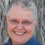 A woman with glasses smiling in front of a tree.