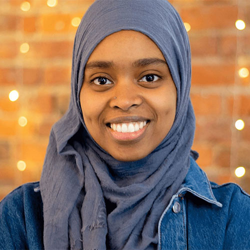 A young woman wearing a hijab smiles in front of a christmas tree.