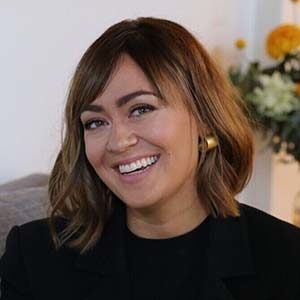 A smiling woman in a black blazer sitting on a couch.