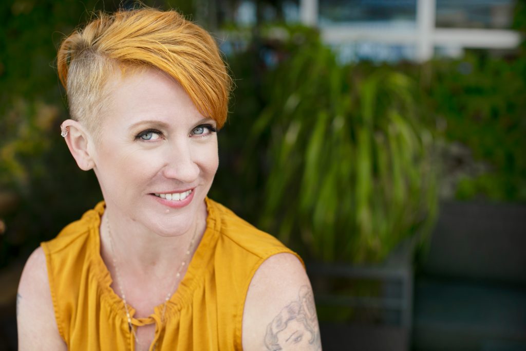 A woman with orange hair and tattoos smiling.