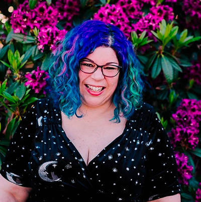 A woman with blue hair and glasses smiles in front of flowers.