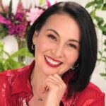 A woman in a red shirt smiling in front of flowers.