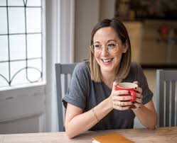 A woman in glasses, smiling, holding a mug at a kitchen table with a notebook beside her.