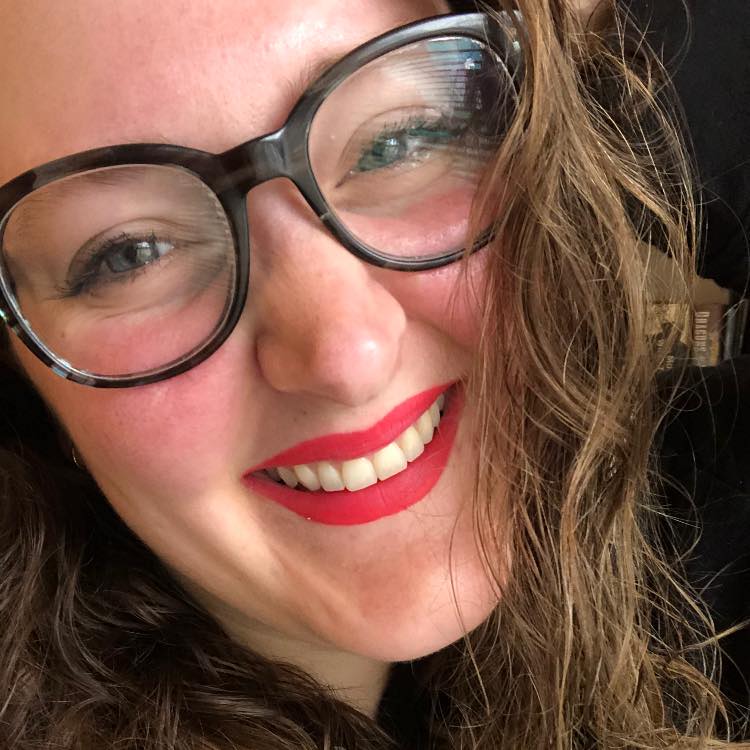 A copywriter with long wavy hair, wearing round glasses and bright red lipstick, smiles closely at the camera.
