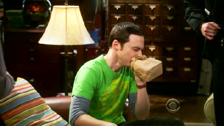 Sheldon sits in a green shirt sits on a couch, breathing rapidly into a brown paper bag, appearing anxious, in a room with a lamp and wooden drawers in the background.